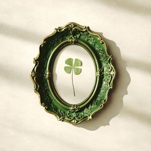 May include: An oval picture frame with an ornate, dark green and gold-toned border featuring raised floral and scrollwork details. Inside the frame, a single, pressed four-leaf clover is centered against a plain, off-white background.