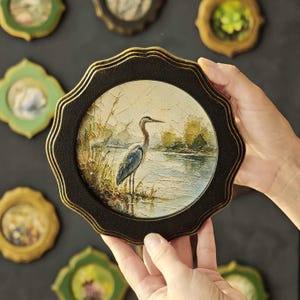 Puede incluir: Pintura redonda enmarcada en negro con borde dorado, que representa una garza en un paisaje pantanoso. La pintura está sostenida por manos, con otras pequeñas obras de arte enmarcadas en el fondo.