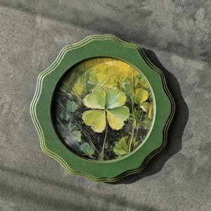 May include: A round, green-framed artwork featuring a four-leaf clover painting. The clover is centered within a field of green and yellow hues, set against a dark background. The frame has a scalloped edge.