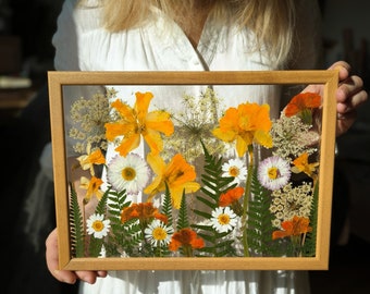 May include: A framed botanical art piece featuring pressed flowers in shades of yellow, orange, white, and green. The wooden frame surrounds a clear panel showcasing the arrangement of various floral elements, including daffodils and daisies.