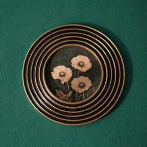 Puede incluir: Una obra de arte circular enmarcada en negro y dorado, que muestra tres flores de amapola crema sobre un fondo verde oscuro. El marco tiene un diseño en capas con círculos concéntricos. La obra de arte está sobre un fondo verde liso.