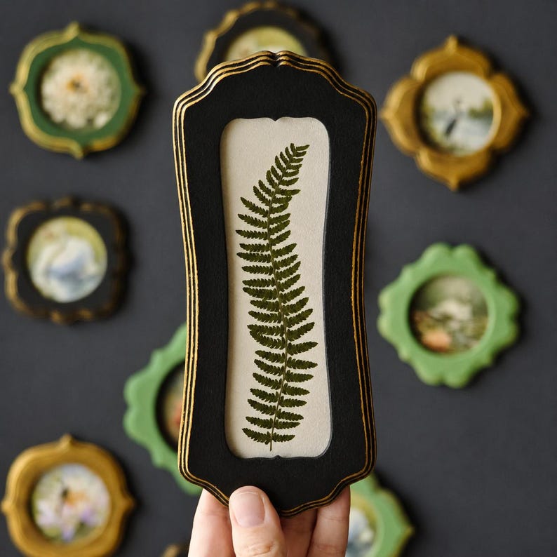 Puede incluir: Un marco rectangular negro y alto con bordes pintados en dorado que contiene una hoja de helecho verde prensada sobre un fondo color crema. El marco presenta una silueta decorativa festoneada y se sostiene verticalmente sobre un fondo gris oscuro.