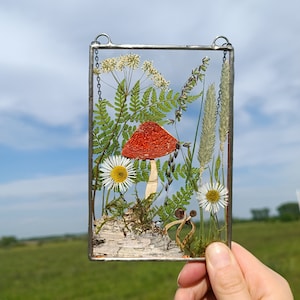 May include: A rectangular glass art piece featuring a red mushroom, white daisies, green ferns, and other dried plants. The artwork is encased in a metal frame with a chain for hanging. The background is a blue sky and green field.