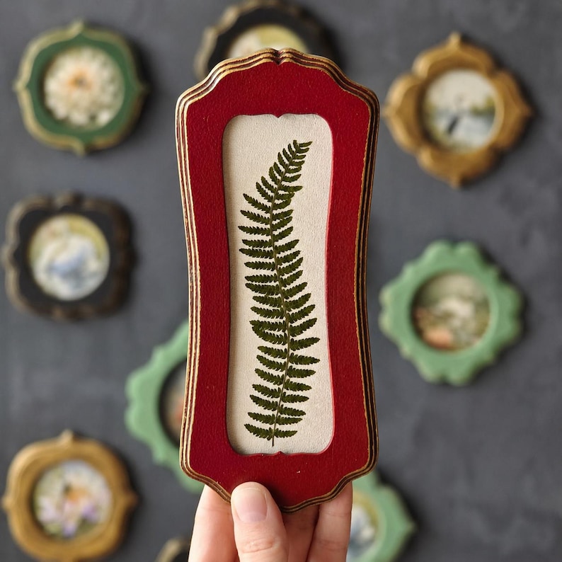 Puede incluir: Un marco de madera rojo, alto y rectangular, con bordes festoneados decorativos, que contiene una hoja de helecho verde prensada centrada sobre un fondo beige claro. El marco presenta un perfil escalonado con textura de madera visible.