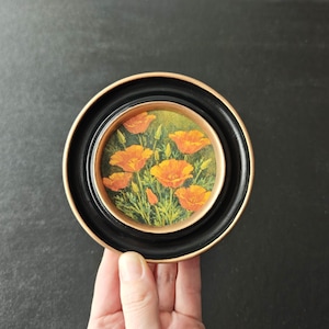 May include: A miniature framed painting of orange poppies on a green backdrop. The artwork is set within a black frame with a gold inner rim. The painting is held by a hand.