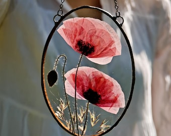 May include: An oval-shaped glass art piece featuring two pressed red poppy flowers with black centers and a seed pod. The flowers are set against a clear background within a black metal frame, suspended by a chain.