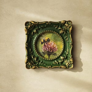 May include: A square wall frame with an ornate, dark green and gold finish surrounding a circular painting. The artwork depicts a bumblebee perched on a textured, pinkish-purple flower against a soft, light green background on a canvas surface.
