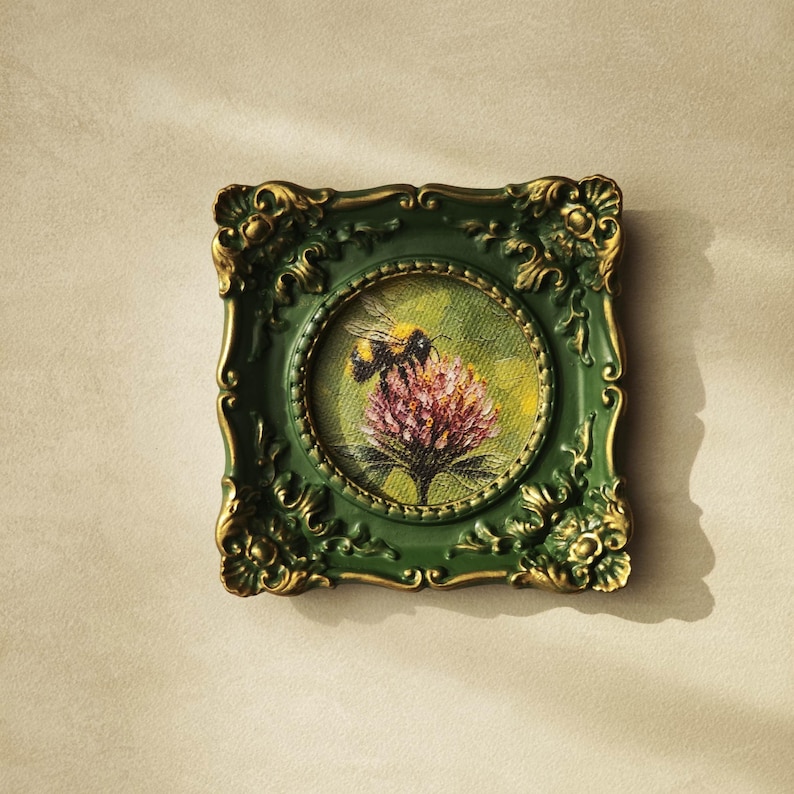 May include: A square wall frame with an ornate, dark green and gold finish surrounding a circular painting. The artwork depicts a bumblebee perched on a textured, pinkish-purple flower against a soft, light green background on a canvas surface.