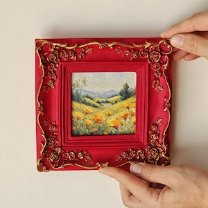 K&ouml;nnte beinhalten: Ein quadratisches Gem&auml;lde einer h&uuml;geligen Landschaft mit gelben Blumen unter einem blassen Himmel, eingefasst in einen leuchtend roten Rahmen mit erhabenen, goldfarbenen Rosenmotiven an den Kanten und Ecken, was f&uuml;r eine strukturierte Optik sorgt.