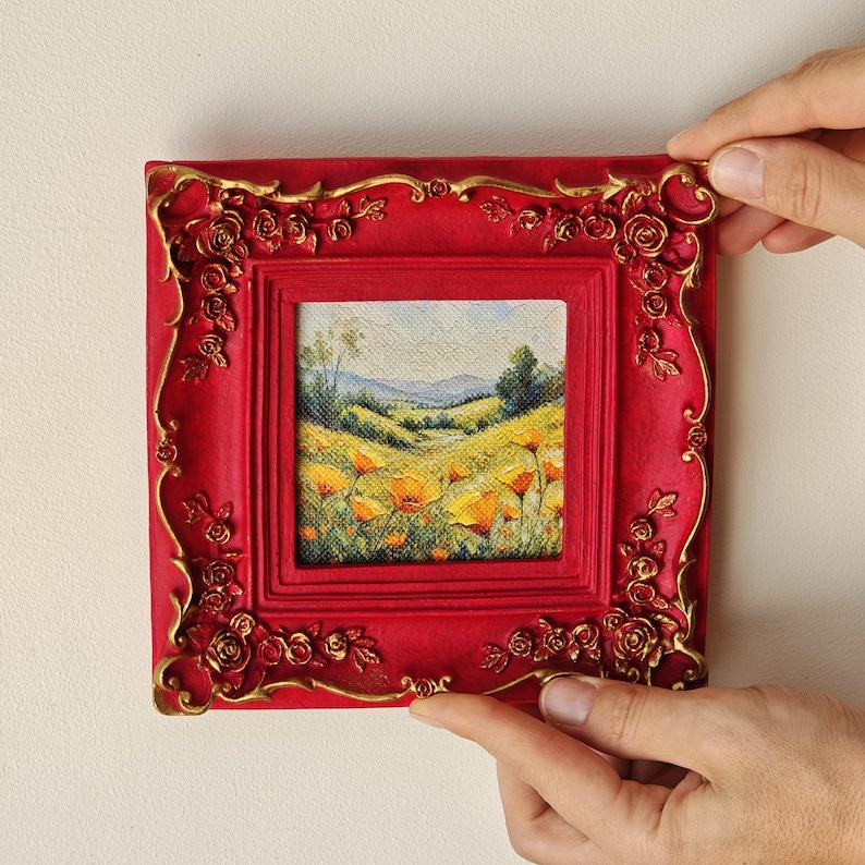 K&ouml;nnte beinhalten: Ein quadratisches Gem&auml;lde einer h&uuml;geligen Landschaft mit gelben Blumen unter einem blassen Himmel, eingefasst in einen leuchtend roten Rahmen mit erhabenen, goldfarbenen Rosenmotiven an den Kanten und Ecken, was f&uuml;r eine strukturierte Optik sorgt.