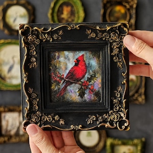 May include: A square wall art piece featuring a vibrant red cardinal bird perched on a branch with red berries. The artwork is encased in a black frame with ornate, raised gold-coloured rose and vine detailing along the edges of the border.