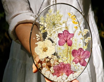 May include: An oval glass frame with dried flowers. The arrangement includes white, yellow, and deep pink blooms, set against a backdrop of delicate greenery. A chain is attached for hanging.