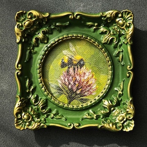 May include: A small square painting in a green and gold ornate frame featuring a bumblebee perched on a pink clover flower. The artwork is rendered in a textured style with a vibrant green background, showcasing the insect and floral details.