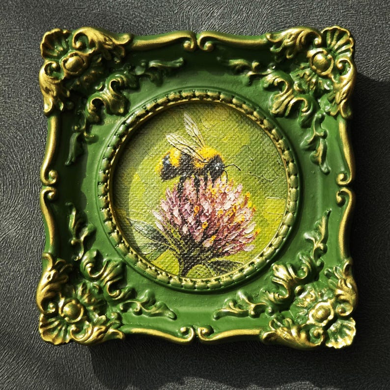 May include: A small square painting in a green and gold ornate frame featuring a bumblebee perched on a pink clover flower. The artwork is rendered in a textured style with a vibrant green background, showcasing the insect and floral details.