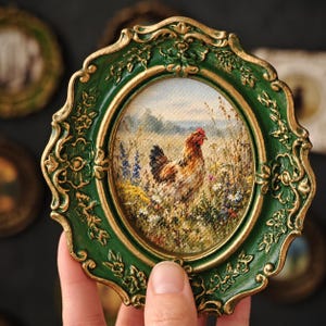 May include: An oval wall art piece featuring a painting of a brown and white chicken standing in a field of wildflowers. The artwork is encased in a decorative, ornate dark green frame with gold-colored highlights along the raised floral edges.