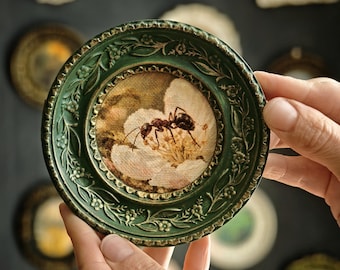 May include: A circular wall art piece featuring a detailed painting of an ant on a white flower. The artwork is encased in a dark green, ornate frame with embossed floral patterns and gold-toned highlights along the edges and inner rim.