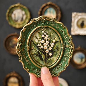 May include: An oval-shaped wall art piece featuring a painting of white lily of the valley flowers with green leaves. The artwork is encased in an ornate, scalloped frame finished in a deep forest green color with gold-toned highlights on the edges.
