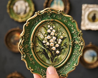 May include: An oval-shaped wall art piece featuring a painting of white lily of the valley flowers with green leaves. The artwork is encased in an ornate, scalloped frame finished in a deep forest green color with gold-toned highlights on the edges.