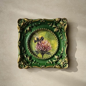 May include: A square, ornate green and gold frame surrounds a circular painting of a bumblebee perched on a pink clover flower. The painting features a textured, yellow-green background, while the frame displays intricate, raised scrollwork details.