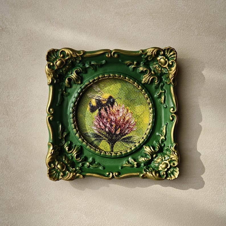 May include: A square, ornate green and gold frame surrounds a circular painting of a bumblebee perched on a pink clover flower. The painting features a textured, yellow-green background, while the frame displays intricate, raised scrollwork details.