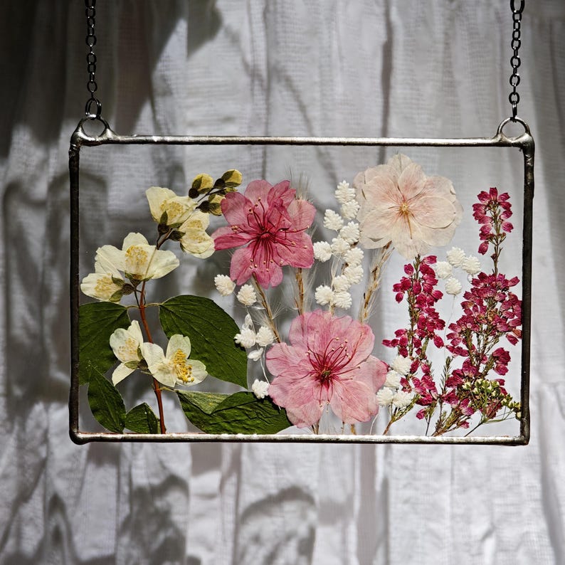 May include: A rectangular glass frame with pressed flowers, including pink, white, and yellow blooms, and green leaves. The frame is suspended by a silver chain, showcasing a botanical arrangement. The flowers are preserved within the clear glass.