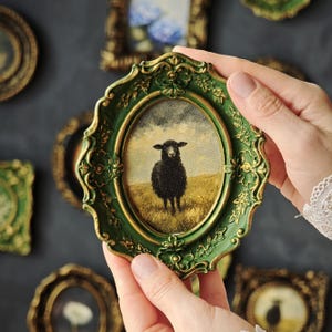 May include: An oval-shaped picture frame with a dark green and gold finish featuring ornate floral relief details. The frame contains a small painting of a black sheep standing in a yellow field under a cloudy sky, held by two hands.