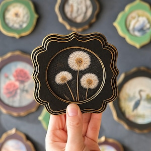 May include: A black, scalloped-edge decorative frame with a gold trim, featuring an embroidered image of three dandelion seed heads. Other frames with floral and nature-themed images are visible in the background.