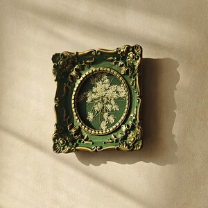 May include: A rectangular wall frame with an ornate, dark green and gold finish featuring intricate scrollwork. The frame contains a central oval display area showcasing a pressed, dried white flower arrangement against a dark green background.