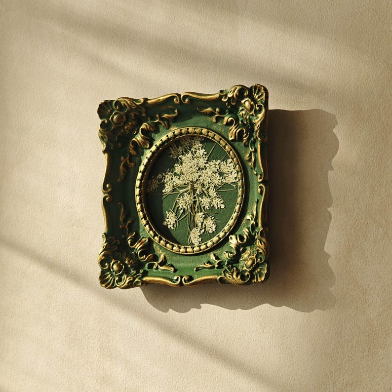 May include: A rectangular wall frame with an ornate, dark green and gold finish featuring intricate scrollwork. The frame contains a central oval display area showcasing a pressed, dried white flower arrangement against a dark green background.