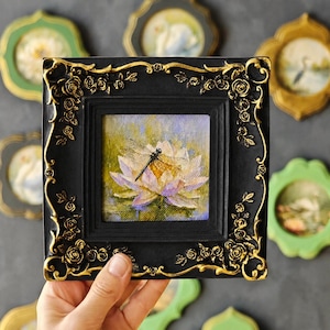 May include: A square painting of a dragonfly resting on a light purple and white water lily, encased in a black frame with ornate gold-colored rose and vine relief detailing along the edges, featuring a textured surface and visible brushstrokes.