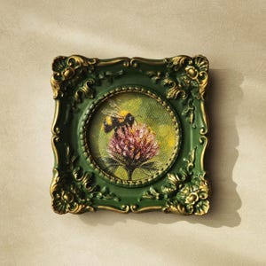 May include: A small square painting featuring a bumblebee on a pink flower, encased in an ornate dark green frame with gold-toned highlights on the decorative relief edges. The artwork is circular within the square frame, showcasing textured brushstrokes.