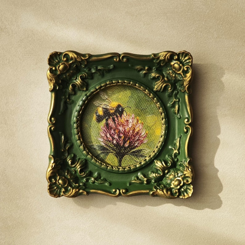May include: A small square painting featuring a bumblebee on a pink flower, encased in an ornate dark green frame with gold-toned highlights on the decorative relief edges. The artwork is circular within the square frame, showcasing textured brushstrokes.