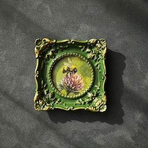 May include: A small square painting featuring a bumblebee perched on a pink flower, enclosed in an ornate green frame with gold-toned highlights. The artwork is circular within the square frame, showcasing detailed brushstrokes and vibrant colors.