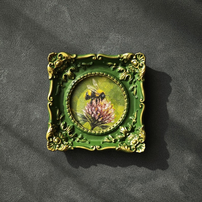 May include: A small square painting featuring a bumblebee perched on a pink flower, enclosed in an ornate green frame with gold-toned highlights. The artwork is circular within the square frame, showcasing detailed brushstrokes and vibrant colors.