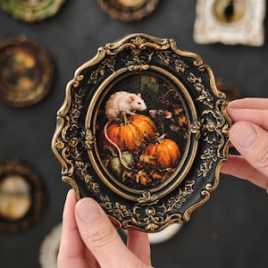 May include: An oval-shaped picture frame with a black and gold finish containing an illustration of a white rat and a dark gray rat sitting among orange pumpkins and autumn leaves. The frame features ornate, raised floral and scrollwork detailing.
