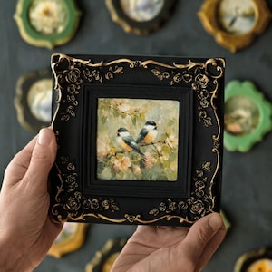 May include: A square painting of two small birds perched on a flowering branch, set within a black frame featuring ornate gold-painted rose relief details. The artwork depicts the birds against a soft, light-coloured, textured floral background.