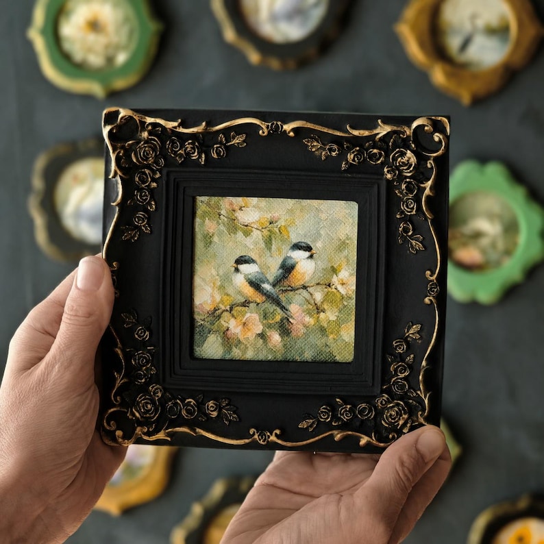 May include: A square painting of two small birds perched on a flowering branch, set within a black frame featuring ornate gold-painted rose relief details. The artwork depicts the birds against a soft, light-coloured, textured floral background.