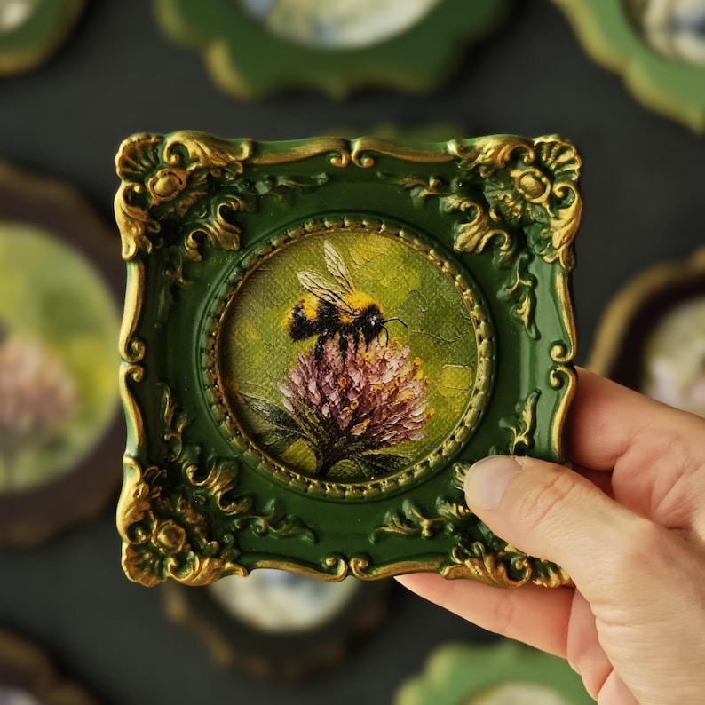 May include: A small square frame in dark green with gold-toned ornate detailing surrounding a circular painting of a bumblebee perched on a pink clover flower. The frame features intricate scrollwork and relief patterns on all four corners.