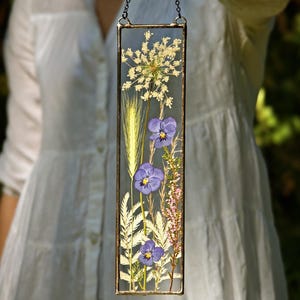 May include: A rectangular glass panel with dried flowers and foliage, including purple pansies, wheat, and delicate white blooms. The panel is framed in a dark metal and suspended by a chain, creating a unique botanical display.