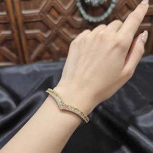May include: A gold bracelet with a V-shaped design, featuring small, sparkling crystals and amber-coloured stones. The bracelet is worn on a wrist, set against a dark, silky background. The bracelet is a stylish piece of jewellery.