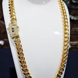Puede incluir: Un collar de cadena de eslabones cubanos en tono dorado grueso con un cierre de caja rectangular. El cierre está incrustado con pequeñas piedras transparentes y muestra un emblema cuadrado negro central con un símbolo dorado en el engaste.