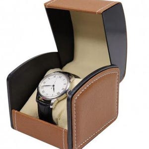 May include: A wristwatch with a white face, black Roman numerals, and a black leather strap displayed inside an open, tan and black storage box. The box features a curved lid, white contrast stitching, and a soft, cream-colored interior cushion.