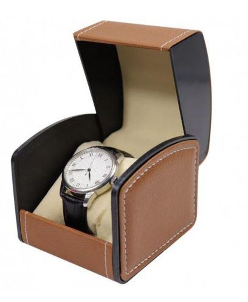 May include: A wristwatch with a white face, black Roman numerals, and a black leather strap displayed inside an open, tan and black storage box. The box features a curved lid, white contrast stitching, and a soft, cream-colored interior cushion.