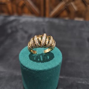 May include: A gold-colored ring with a unique design. The ring features a series of curved, ribbed bands, each adorned with small, sparkling stones. The ring is displayed on a teal green cylindrical stand, with a dark background.