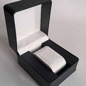 May include: A rectangular watch display box with a textured black exterior and a hinged lid. The interior features a white satin-lined lid and a removable white velvet-textured watch cushion centered within a black base compartment.