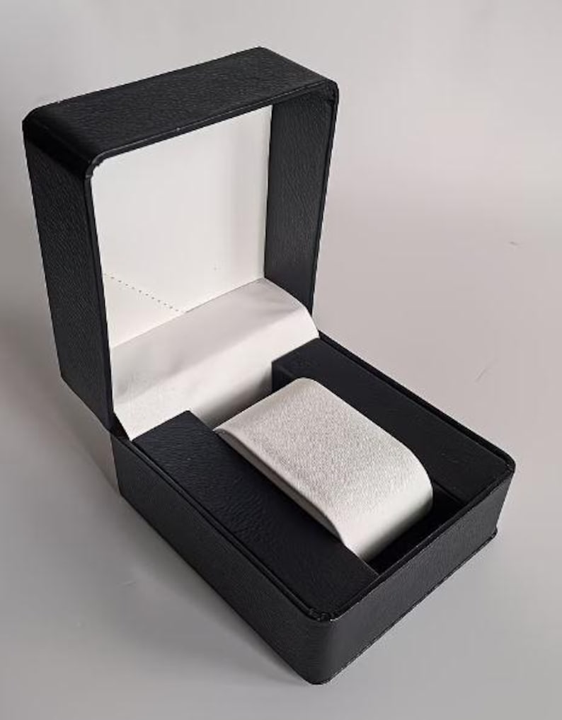 May include: A rectangular watch display box with a textured black exterior and a hinged lid. The interior features a white satin-lined lid and a removable white velvet-textured watch cushion centered within a black base compartment.