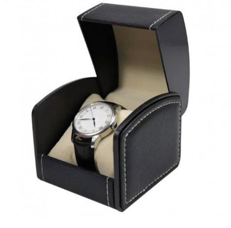 May include: A black leatherette watch box with white contrast stitching, featuring an open lid and a cream-colored interior cushion. Inside, a silver-toned wristwatch with a white face and a black leather strap is displayed on the cushion.