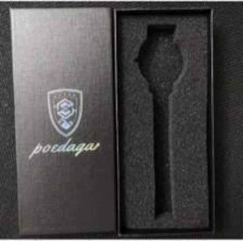 May include: A black rectangular watch box with a textured finish. The lid features a silver shield logo with the text poedaga below it. The interior contains a black foam insert with a cutout shaped to hold a wristwatch securely in place.