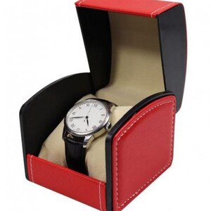 May include: A wristwatch with a white face, black Roman numerals, and a black leather strap, displayed inside an open, red and black storage box with white stitching and a beige interior lining designed to hold the timepiece securely.