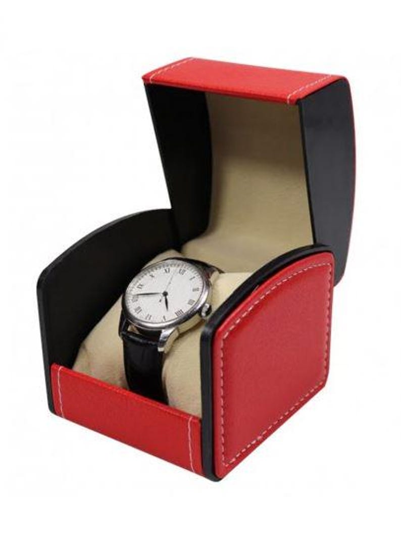 May include: A wristwatch with a white face, black Roman numerals, and a black leather strap, displayed inside an open, red and black storage box with white stitching and a beige interior lining designed to hold the timepiece securely.